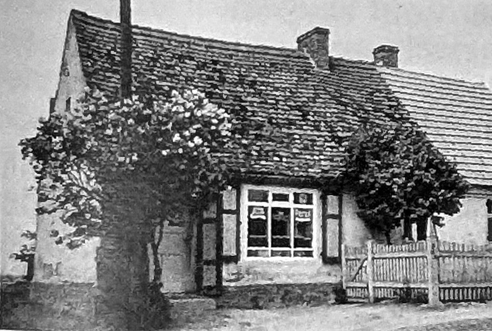 Schwarzweiß-Aufnahme: ein niedriges Haus mit Ziegeldach, davor ein blühender Strauch und ein Lattenzaun