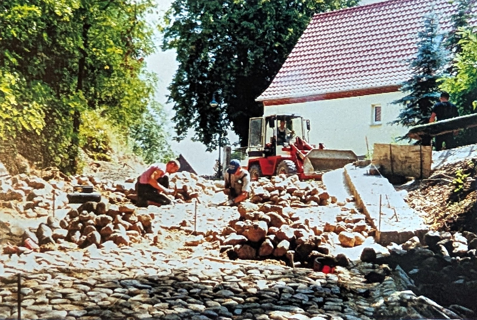 Farbfoto: Männer setzen Feldsteine in einen Hohlweg, dahinter ein Radlader und ein Haus mit rotem Dach